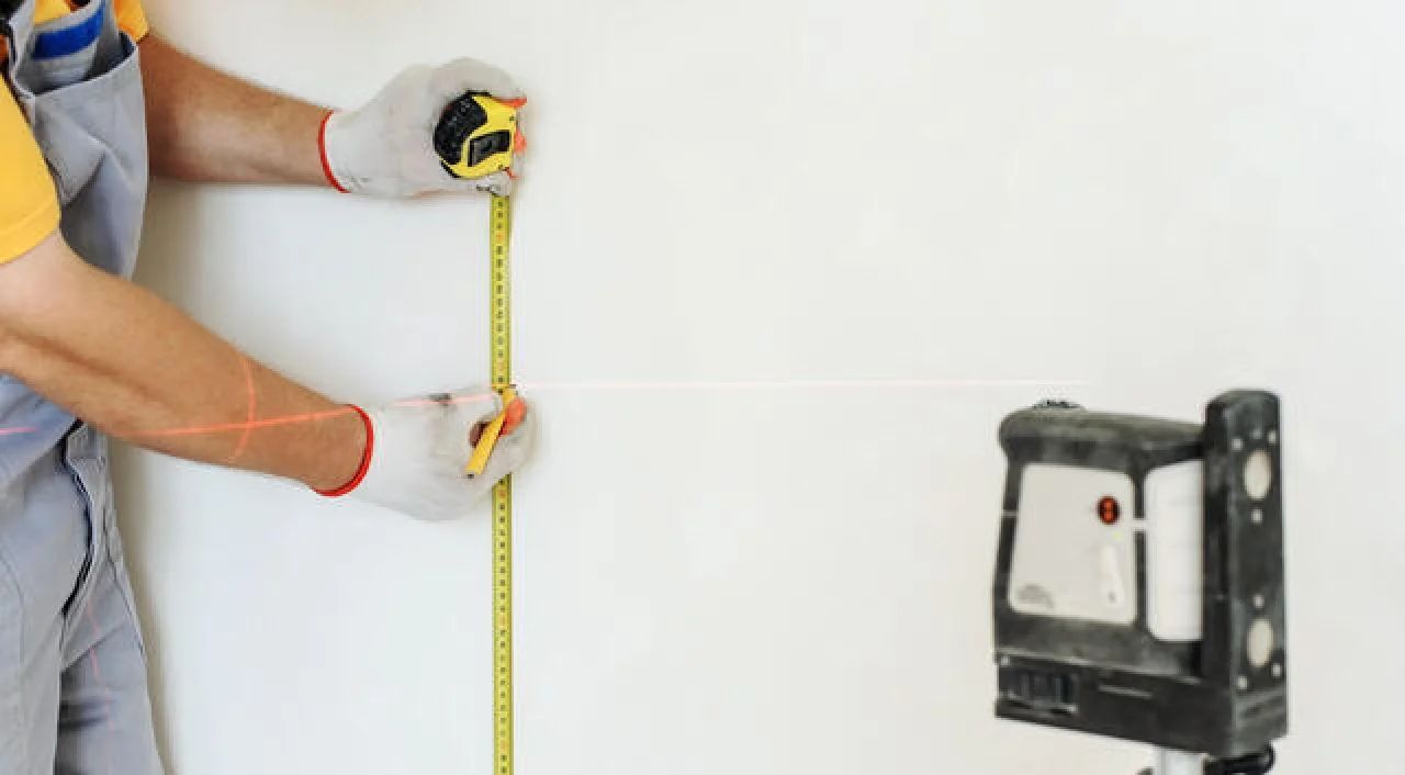 Person in gloves measuring a white wall with a tape measure near a mounted tool bracket
