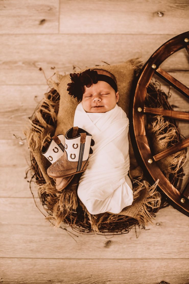A newborn baby is wrapped in a white blanket and holding a pair of cowboy boots.