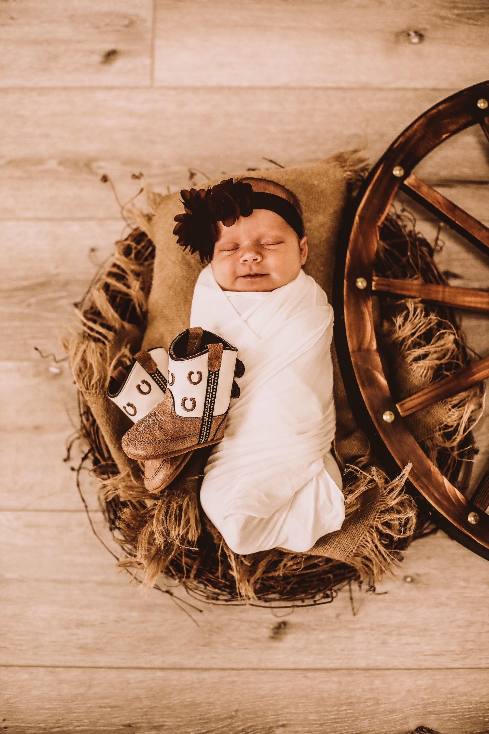 A newborn baby is wrapped in a white blanket and holding a pair of cowboy boots.
