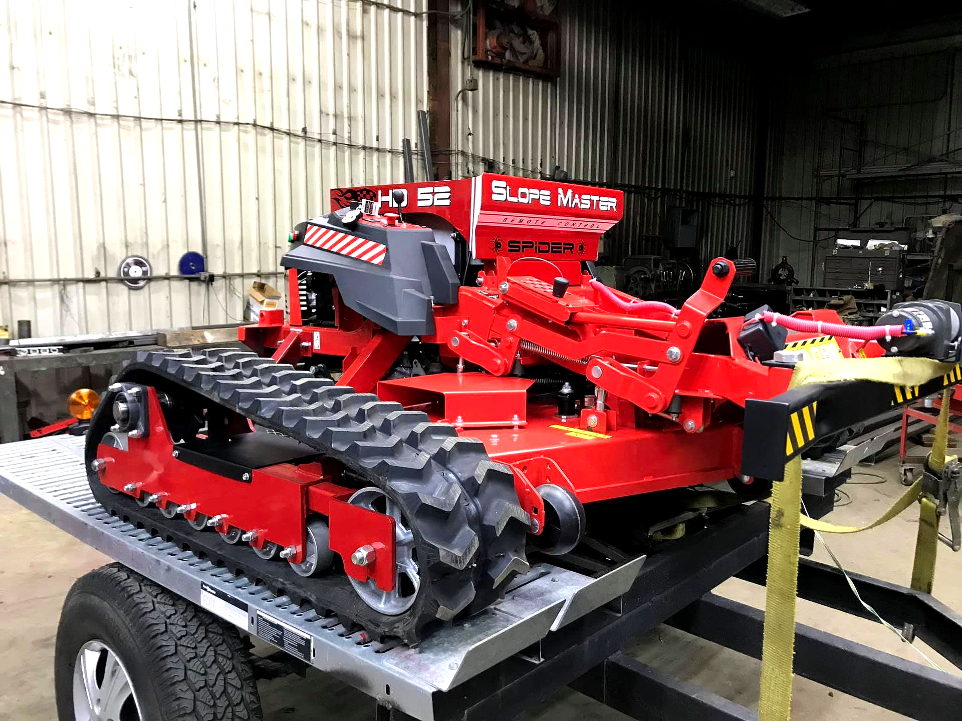 Our Models Red tracked Slope Master Innovations Slope Mower machine with a red hopper on a flatbed trailer inside a warehouse, clearly showing the commercial-grade, tank-like tracks.