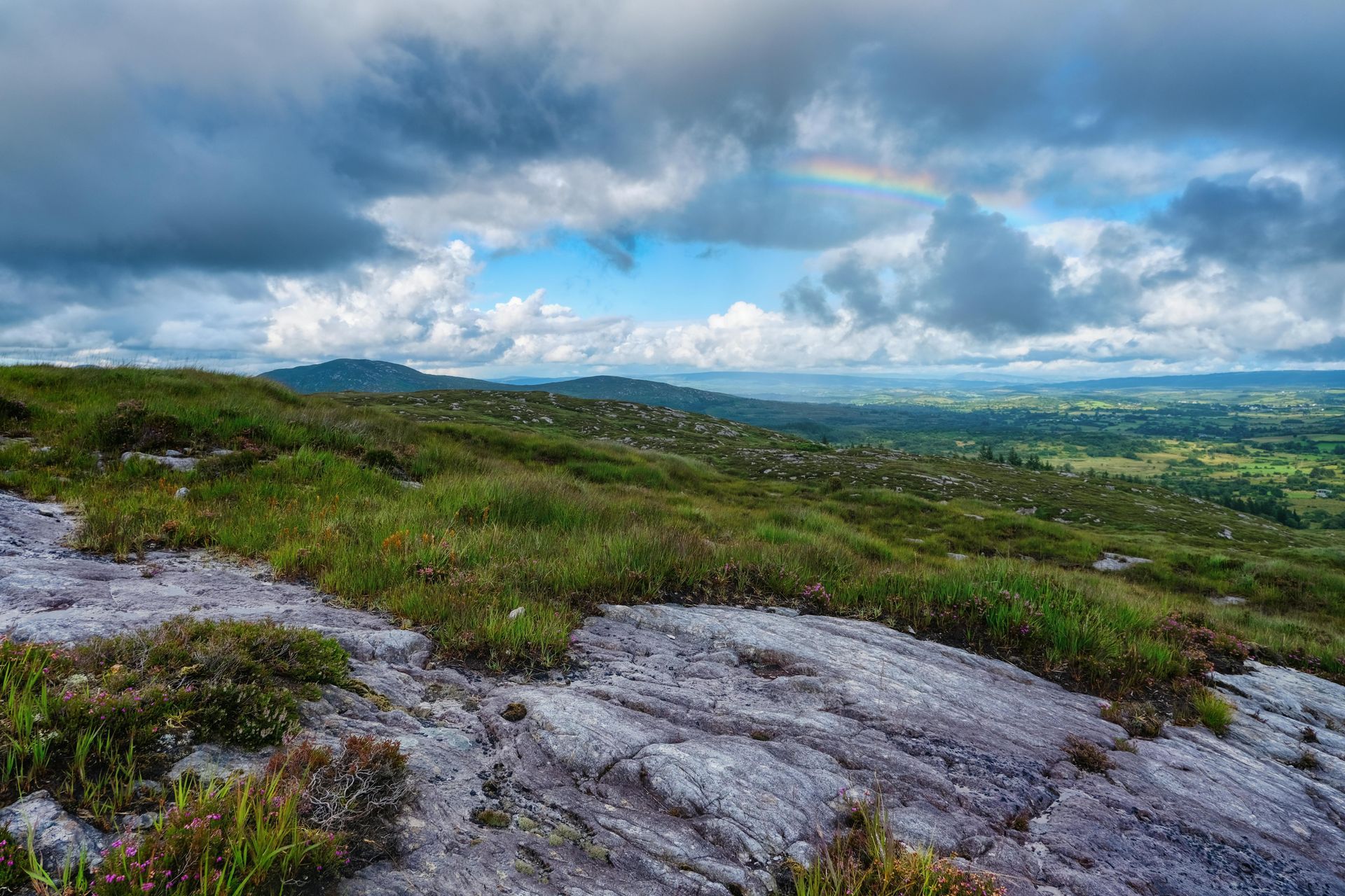 Rocky hillside with green vegetation, rainbow, and distant valley under a cloudy sky.