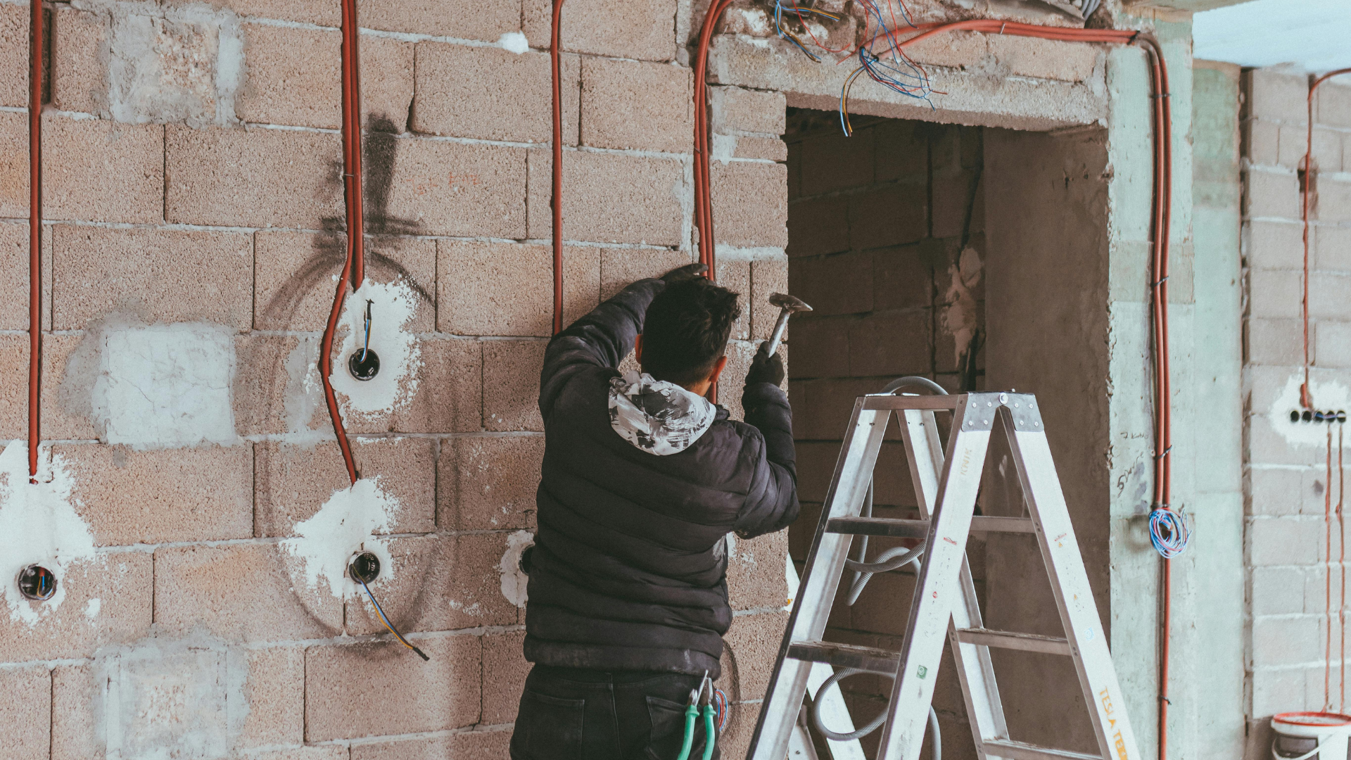 Construction worker hammering near electrical wiring on a cinder block wall.