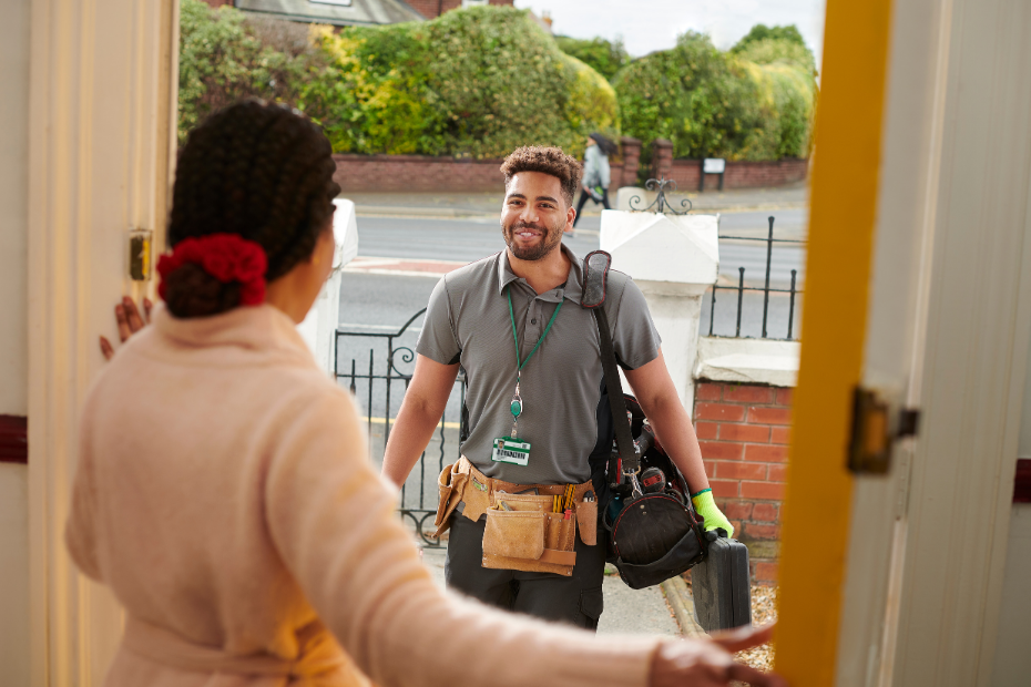 A woman opens a door for a smiling repairman holding tools, standing on a brick walkway outside a house.