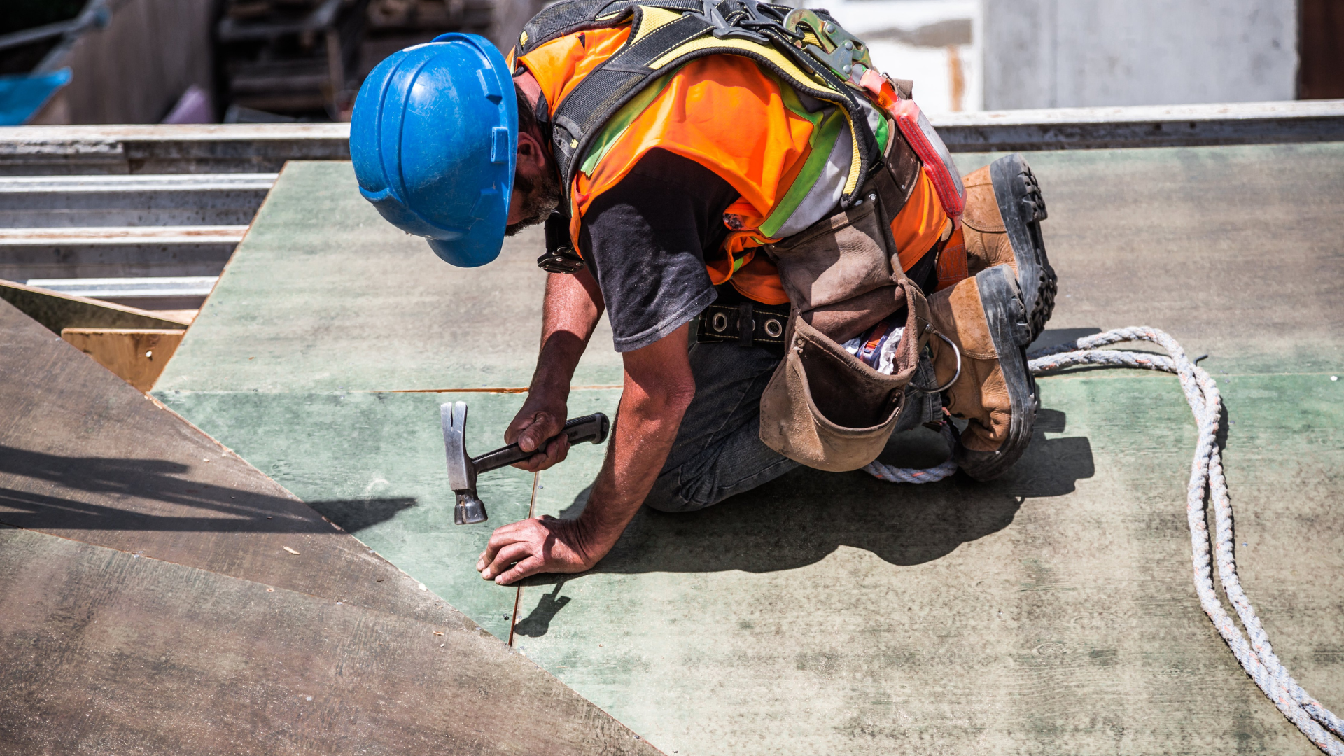 Construction worker on a roof hammering, wearing a blue hard hat, orange vest, and safety harness.
