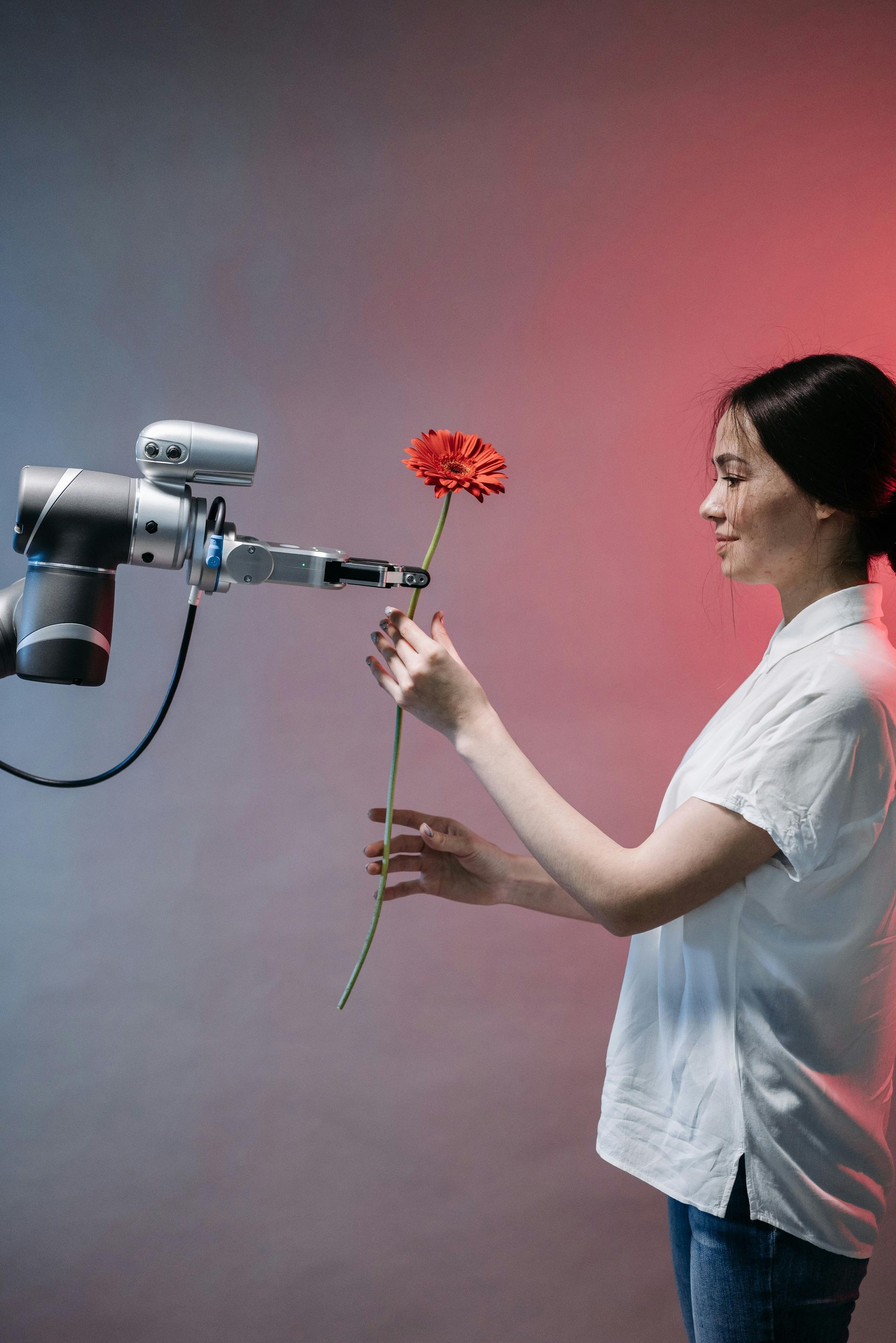 A woman is holding a red flower in front of a robotic arm.