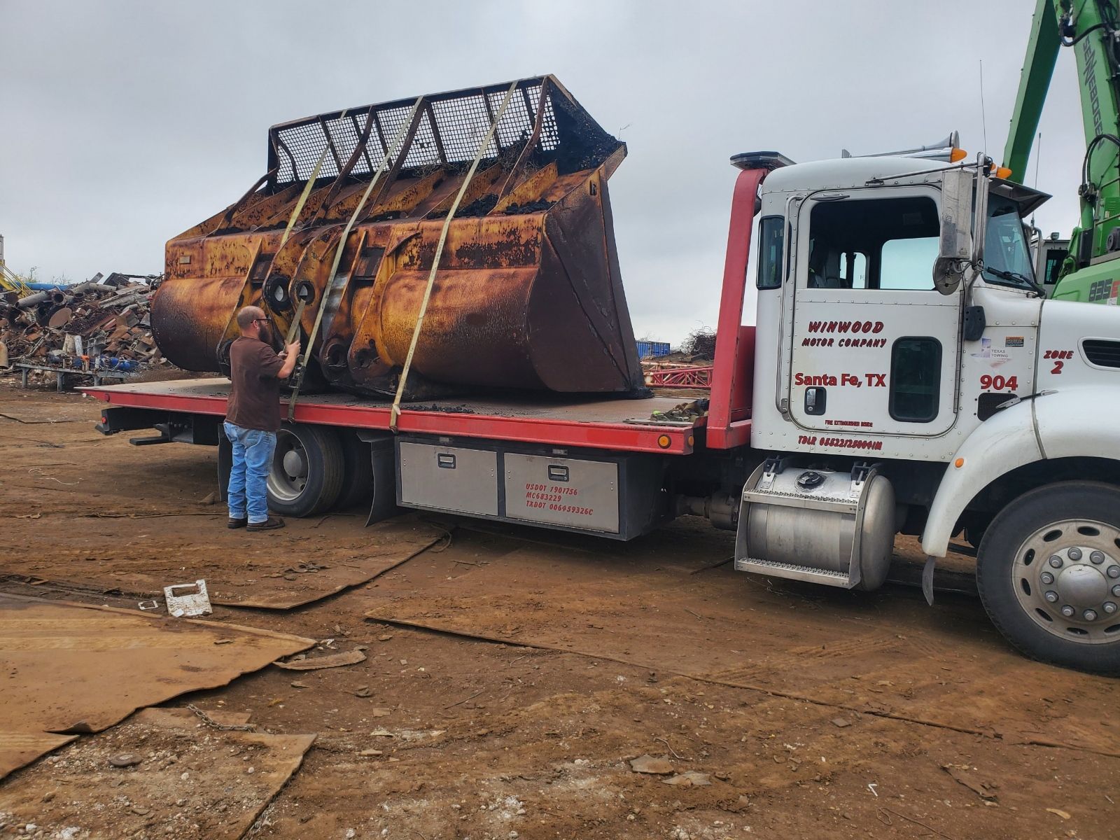 A large rusty excavator bucket is secured on a flatbed truck at a salvage yard. A person secures straps.