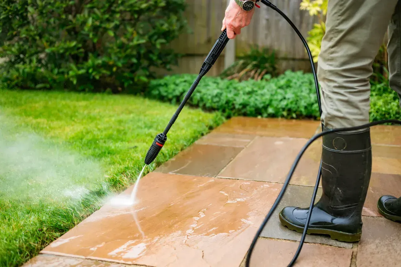 Person power washing a stone patio with a high-pressure hose in a backyard, surrounded by grass and plants.
