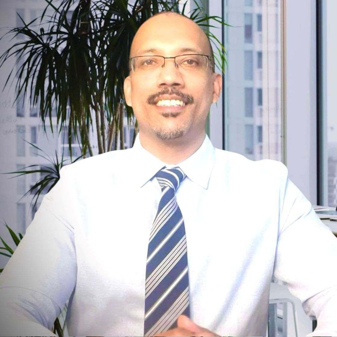 Smiling man in a white shirt and striped tie seated in an office with plants and windows behind him