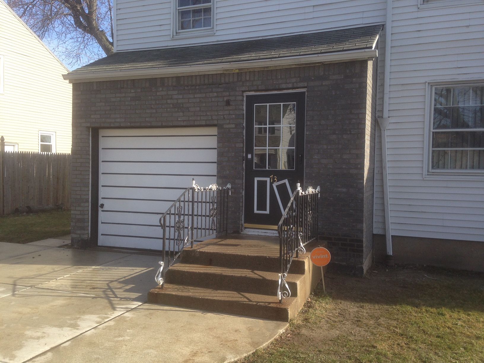 Garage and entry with steps, gray brick, white garage door, black door. Curving metal railing, orange light.