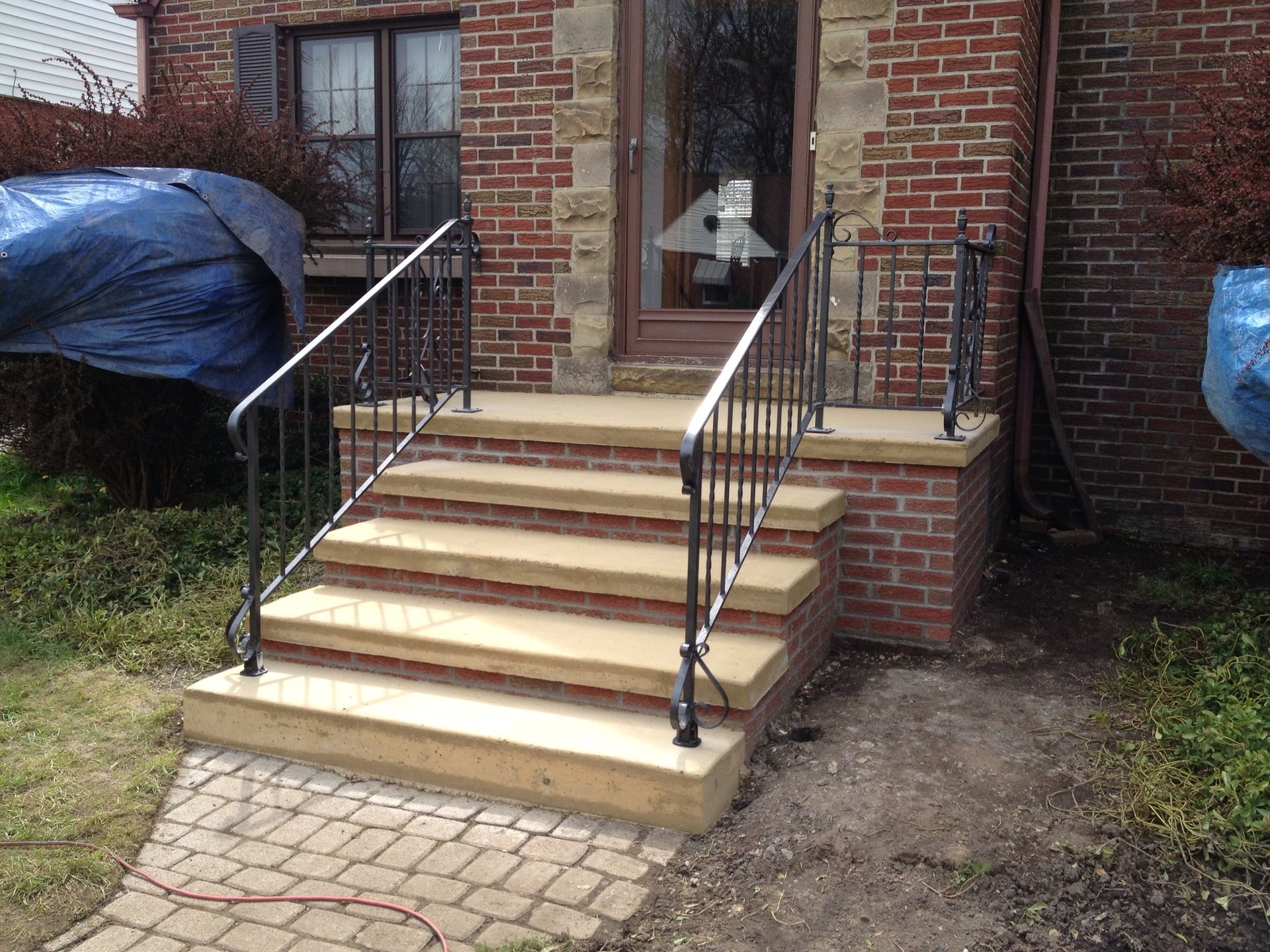 Brick home with concrete steps and black iron railings; a blue tarp is visible on the left.