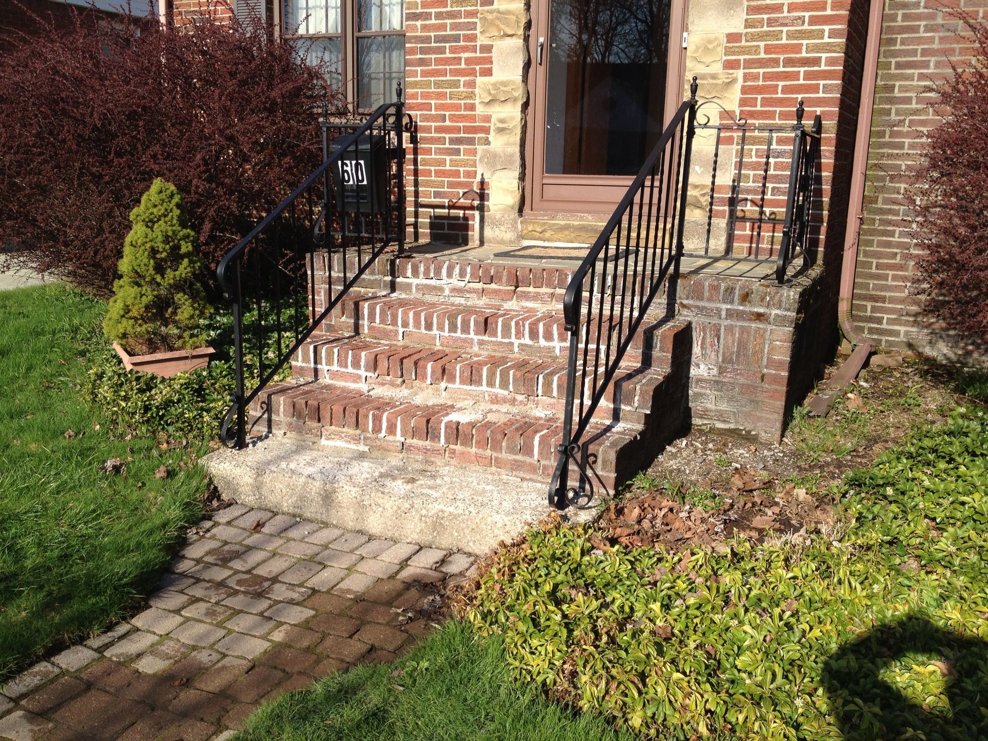 Brick steps leading to a house entrance with black metal railings. Green grass and shrubs surround.