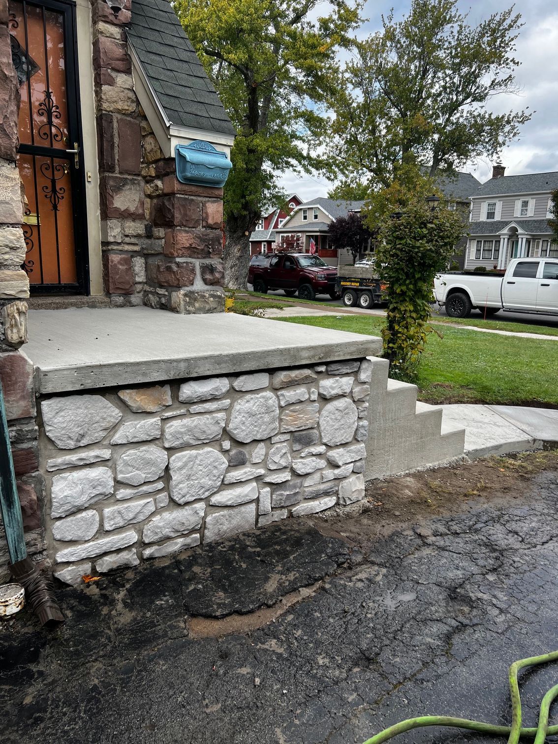 Stone front steps and porch beside a brick house, with a sidewalk, lawn, and parked trucks on a residential street.
