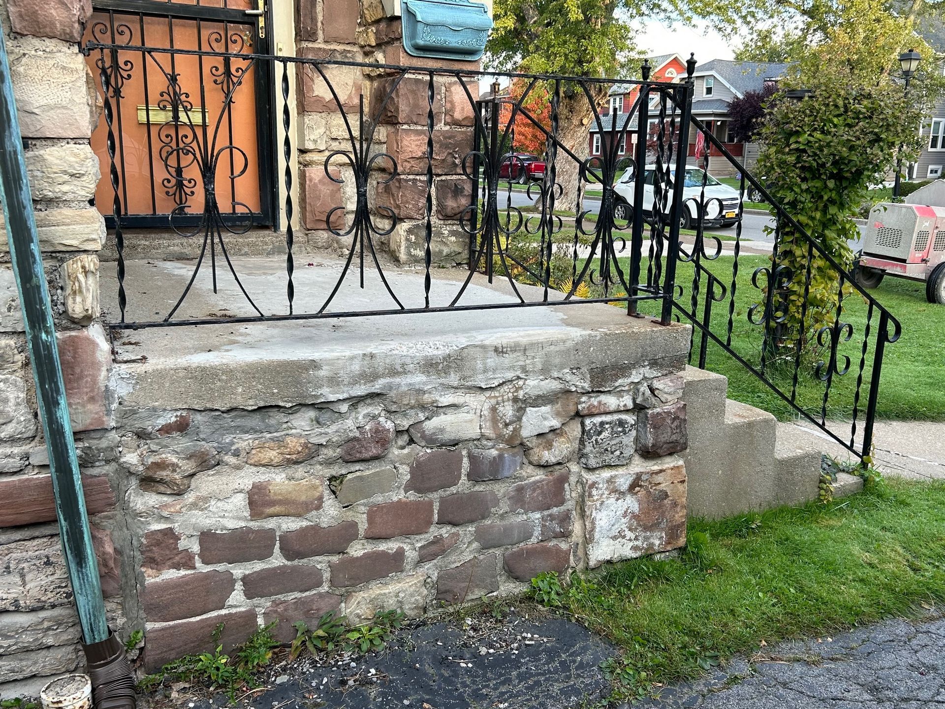 Stone front steps and porch with black wrought-iron railing, bordered by grass and a sidewalk.