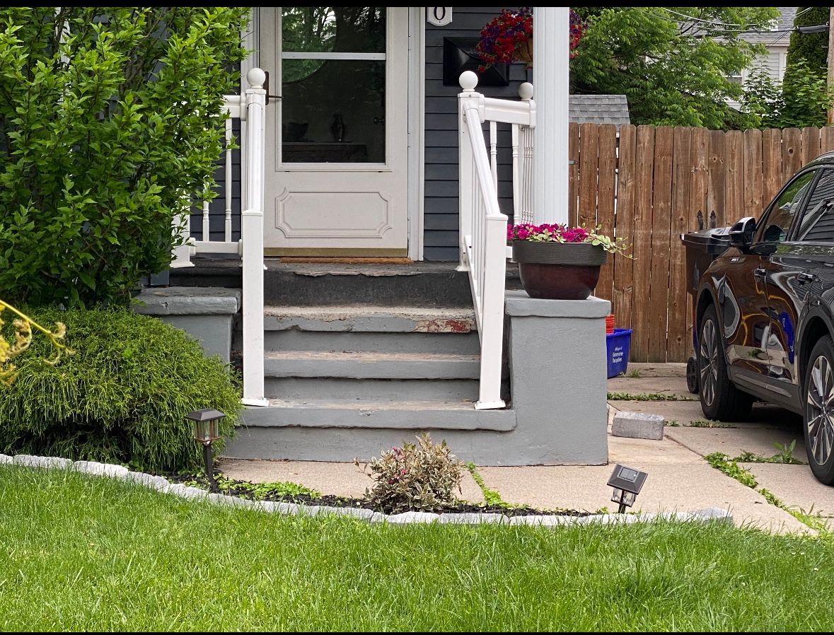 Front porch with white railings, steps, potted flowers, and a parked car beside a lawn.