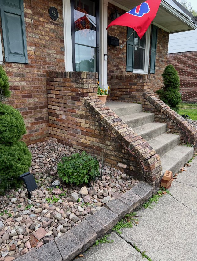 Brick front porch with steps, brick walls, and two red flags beside a small landscaped garden.