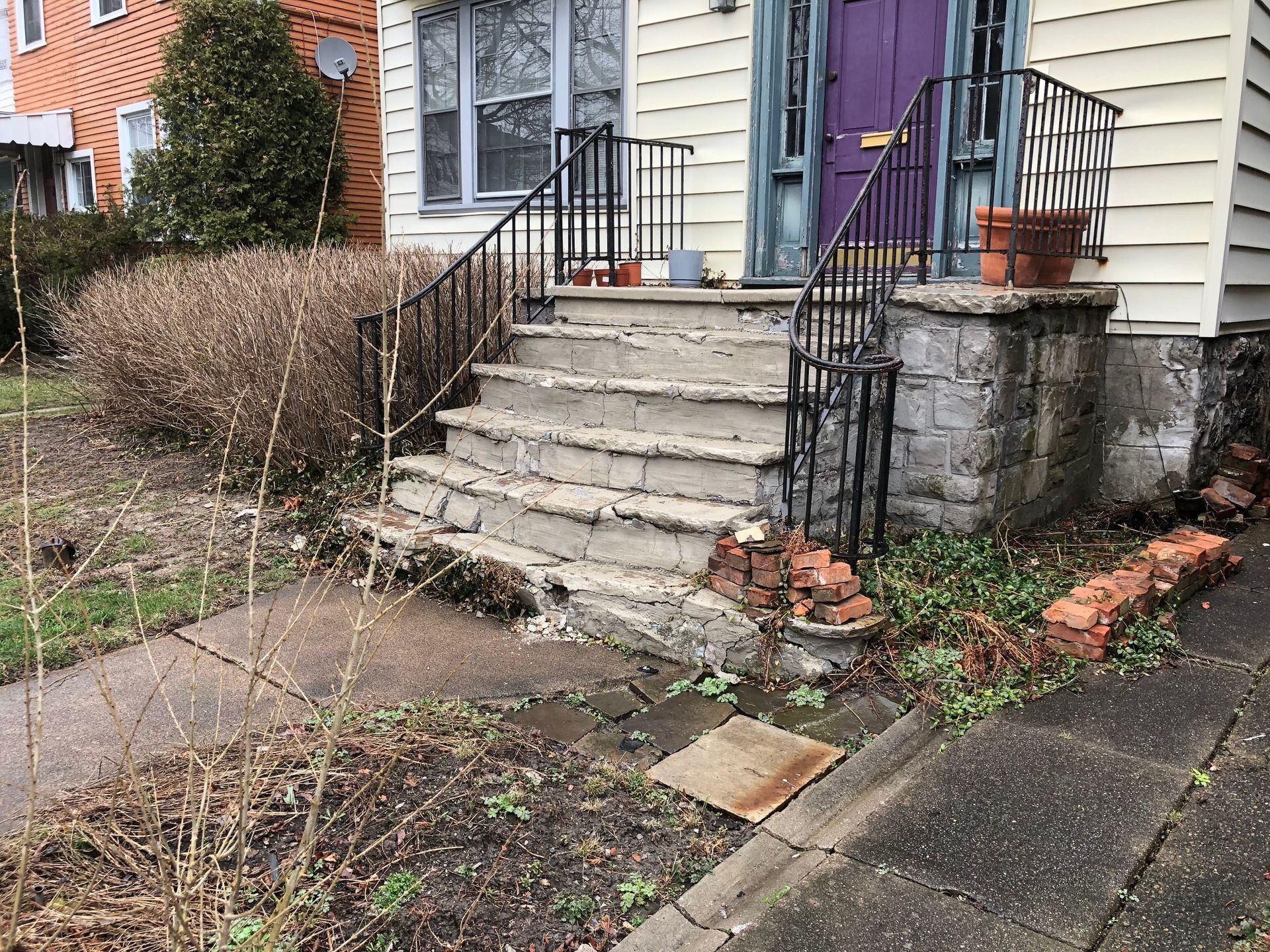 Front steps with cracked concrete and black railings leading to a purple front door beside a house and sidewalk.