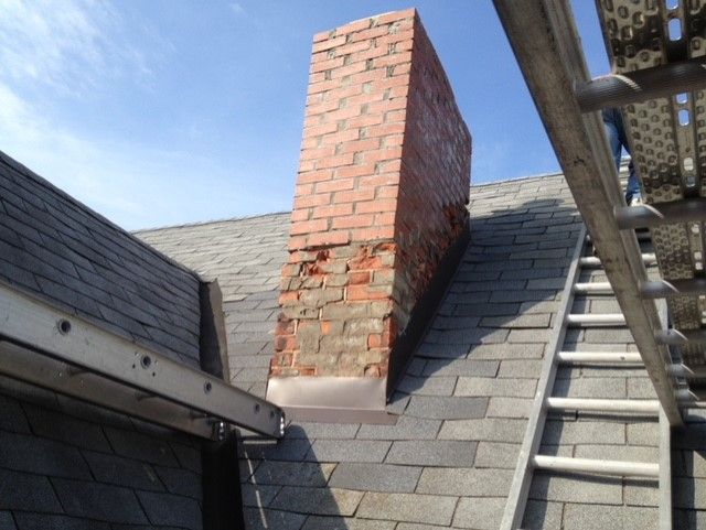 Brick chimney on a dark roof, showing significant brick damage and deterioration.