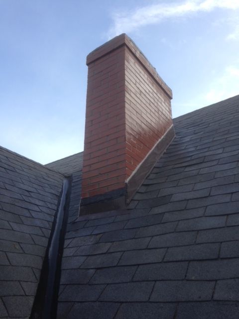 Brick chimney on a dark roof against a cloudy sky, with branches overhead.