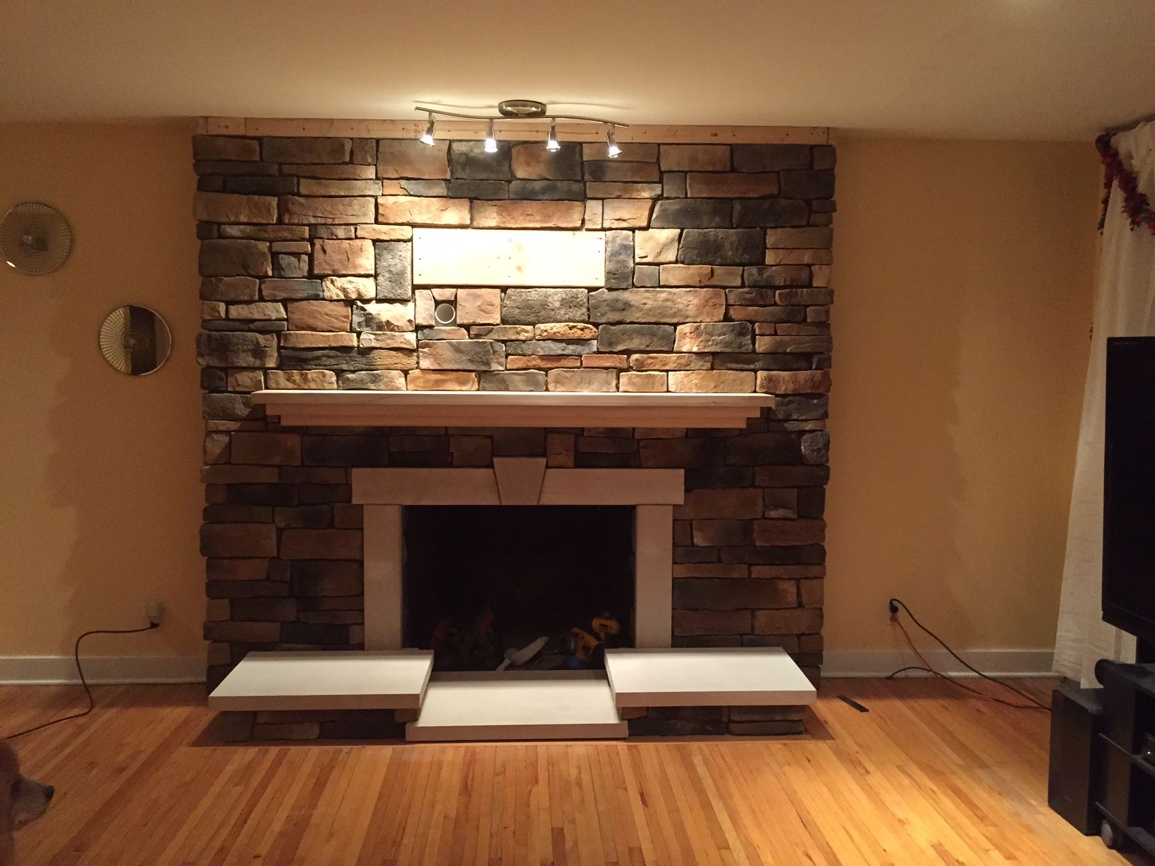 Stone fireplace with light wooden mantel, inset hearth, and three overhead spotlights.