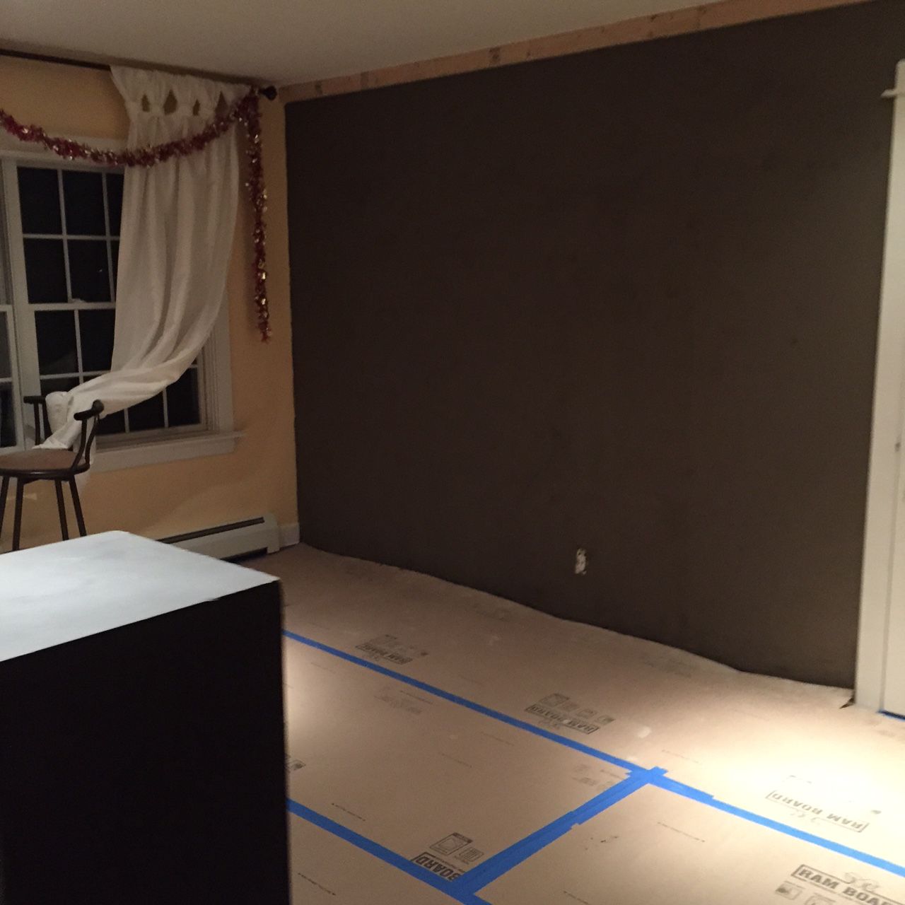 Room with dark brown accent wall, window with white curtain and red garland. Floor covered in cardboard.