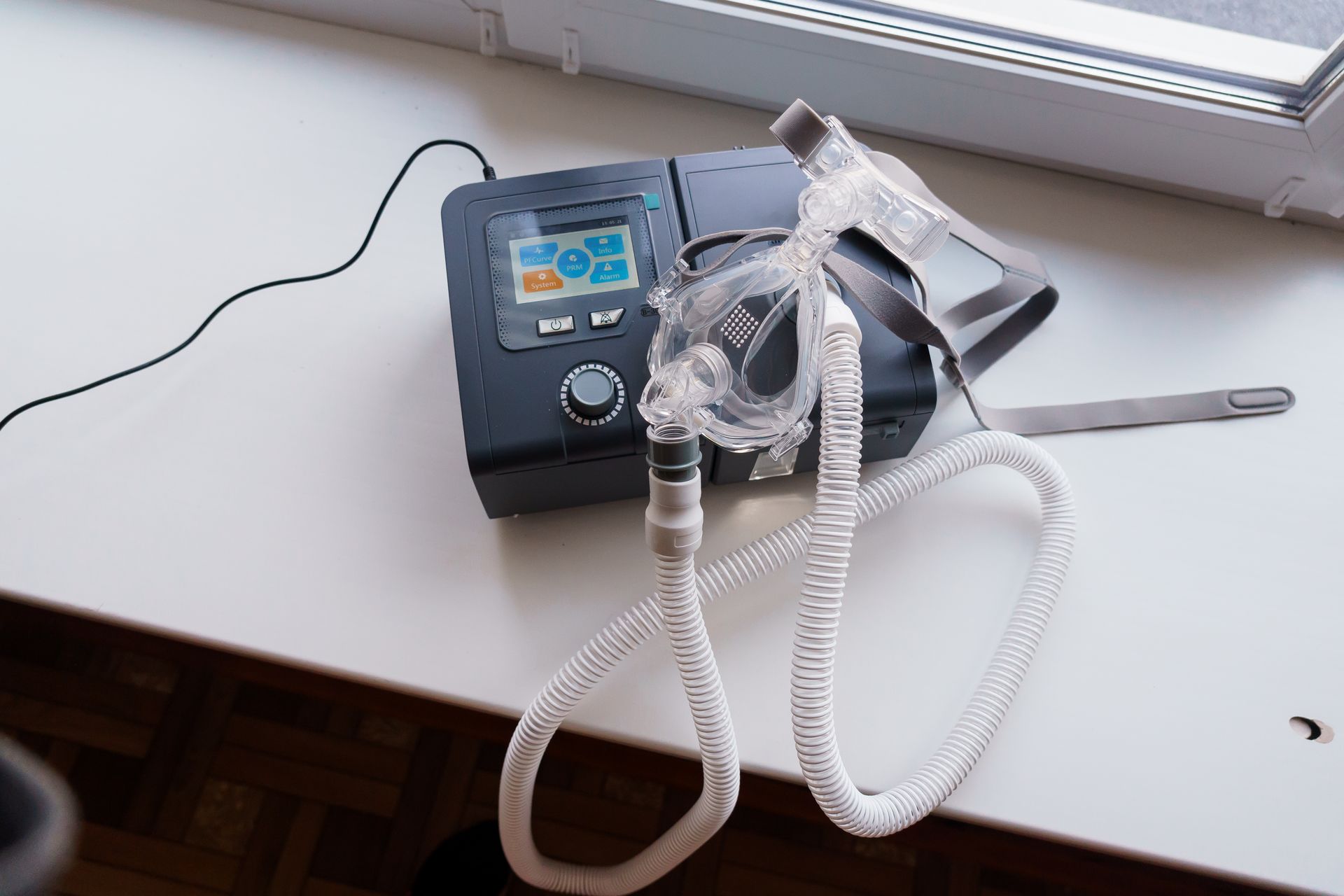 CPAP machine with coiled breathing tubes on a white tabletop near a window.