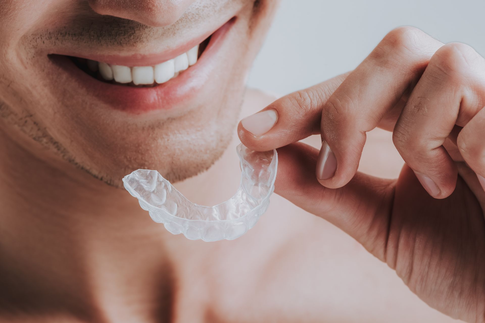Person smiling while holding a clear dental aligner near their mouth.