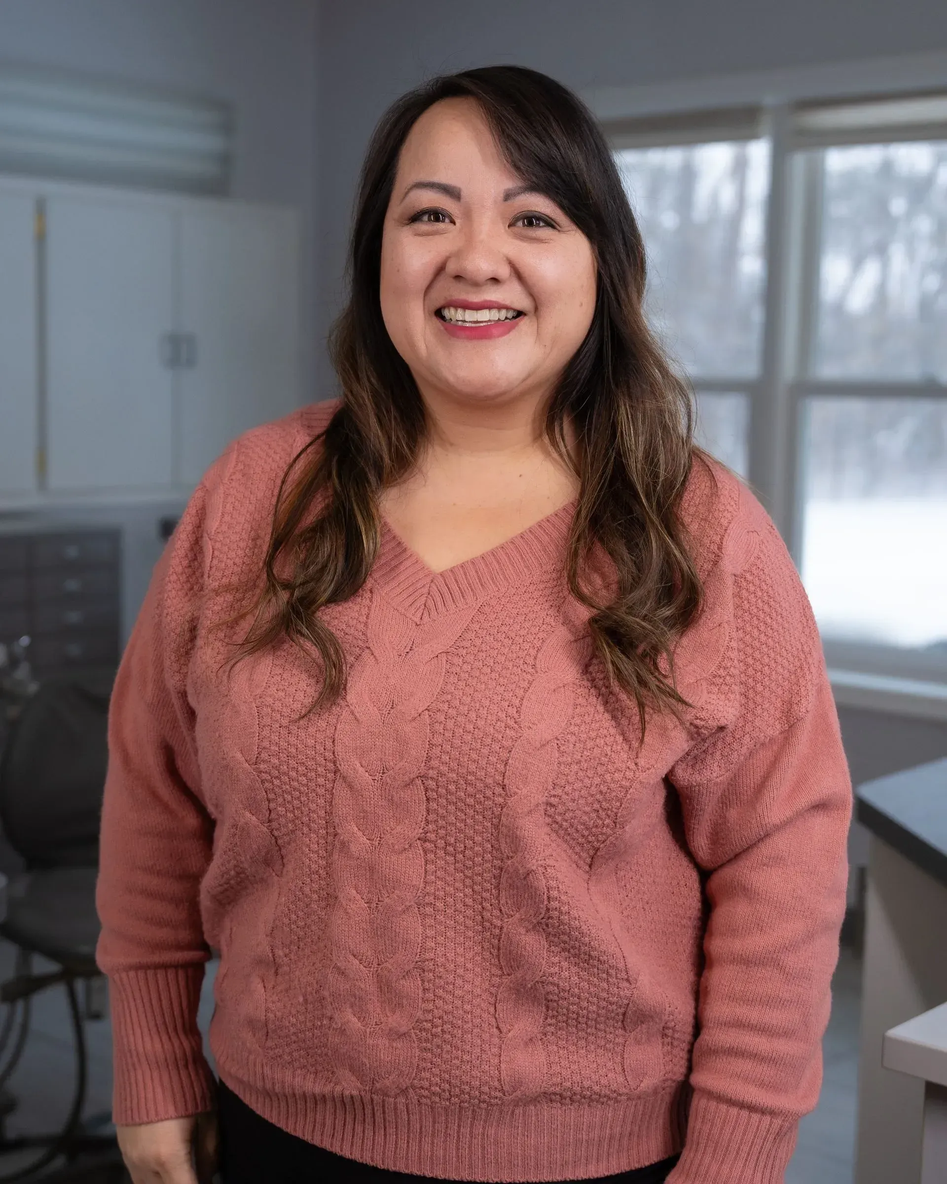 Smiling person in a pink sweater standing in a bright room with large windows.
