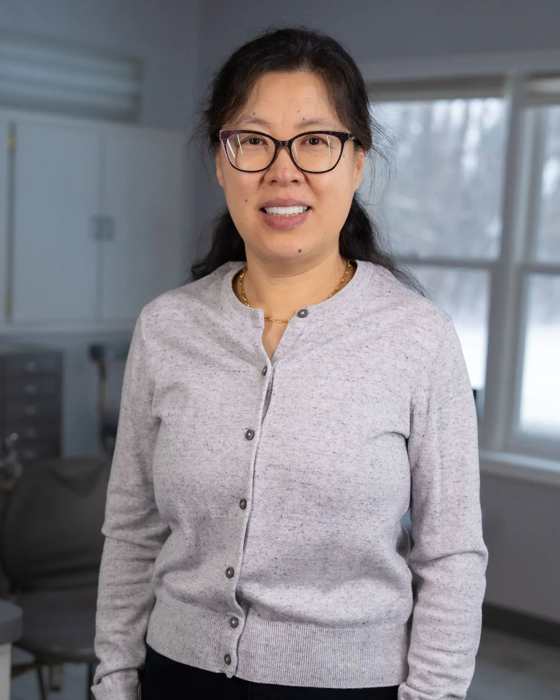 Person wearing glasses and a light gray sweater stands indoors near windows, smiling at the camera.