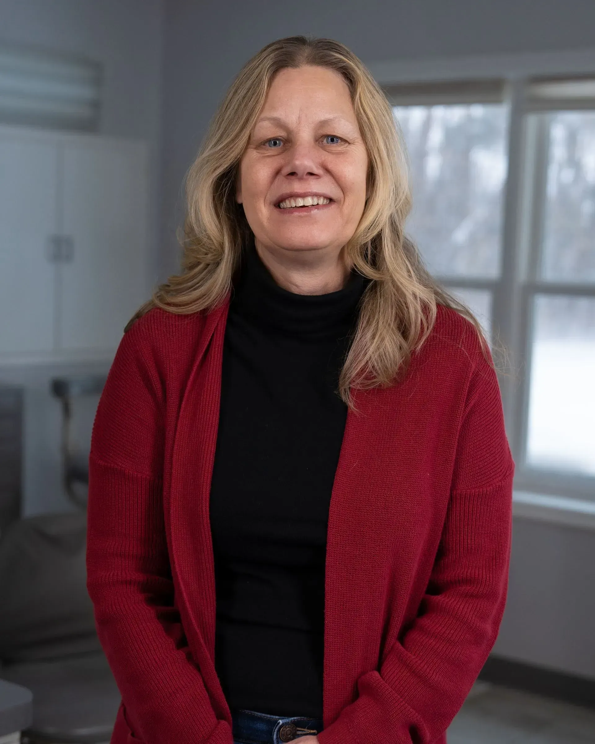 Smiling woman in a red cardigan standing indoors near a window.