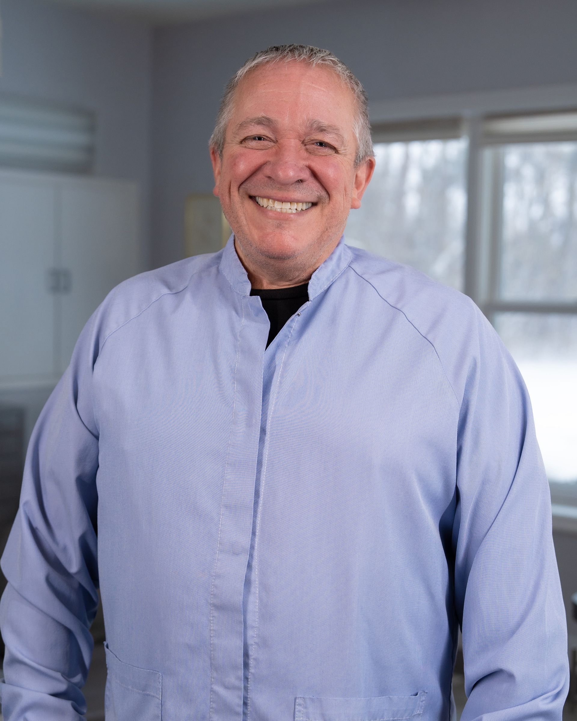 Smiling man in a light blue shirt standing in a bright room with windows behind him.
