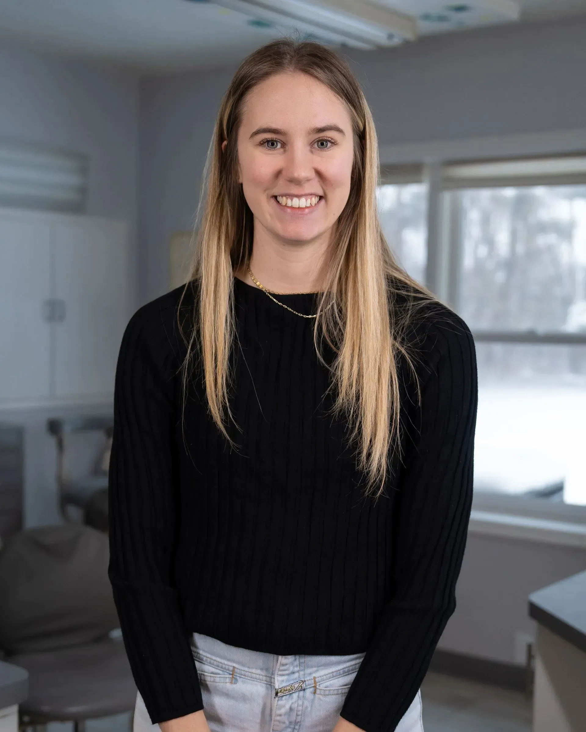 Smiling woman in a black sweater standing indoors near large windows and kitchen counters.