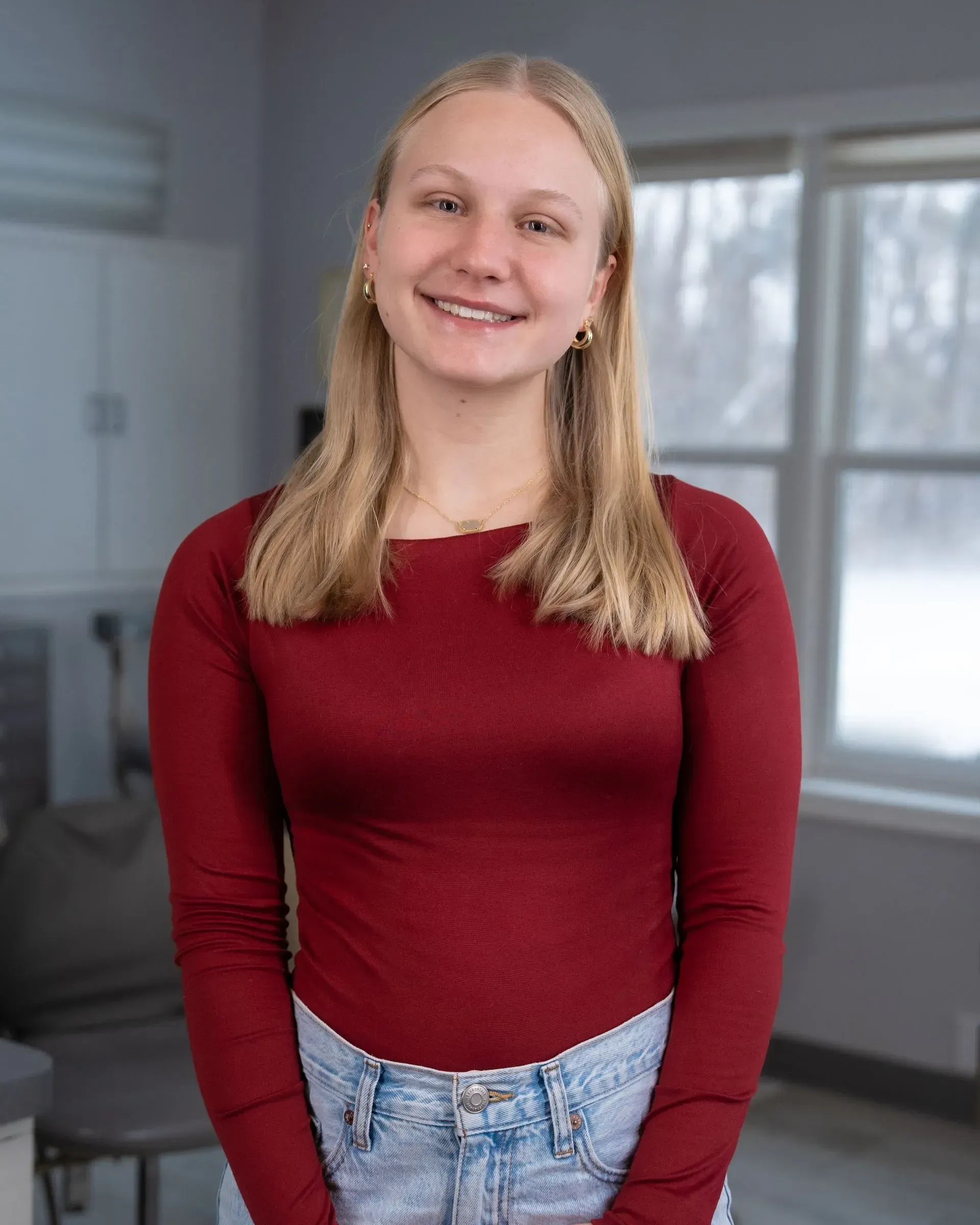 Person smiling in a red top and jeans, standing indoors near a window.
