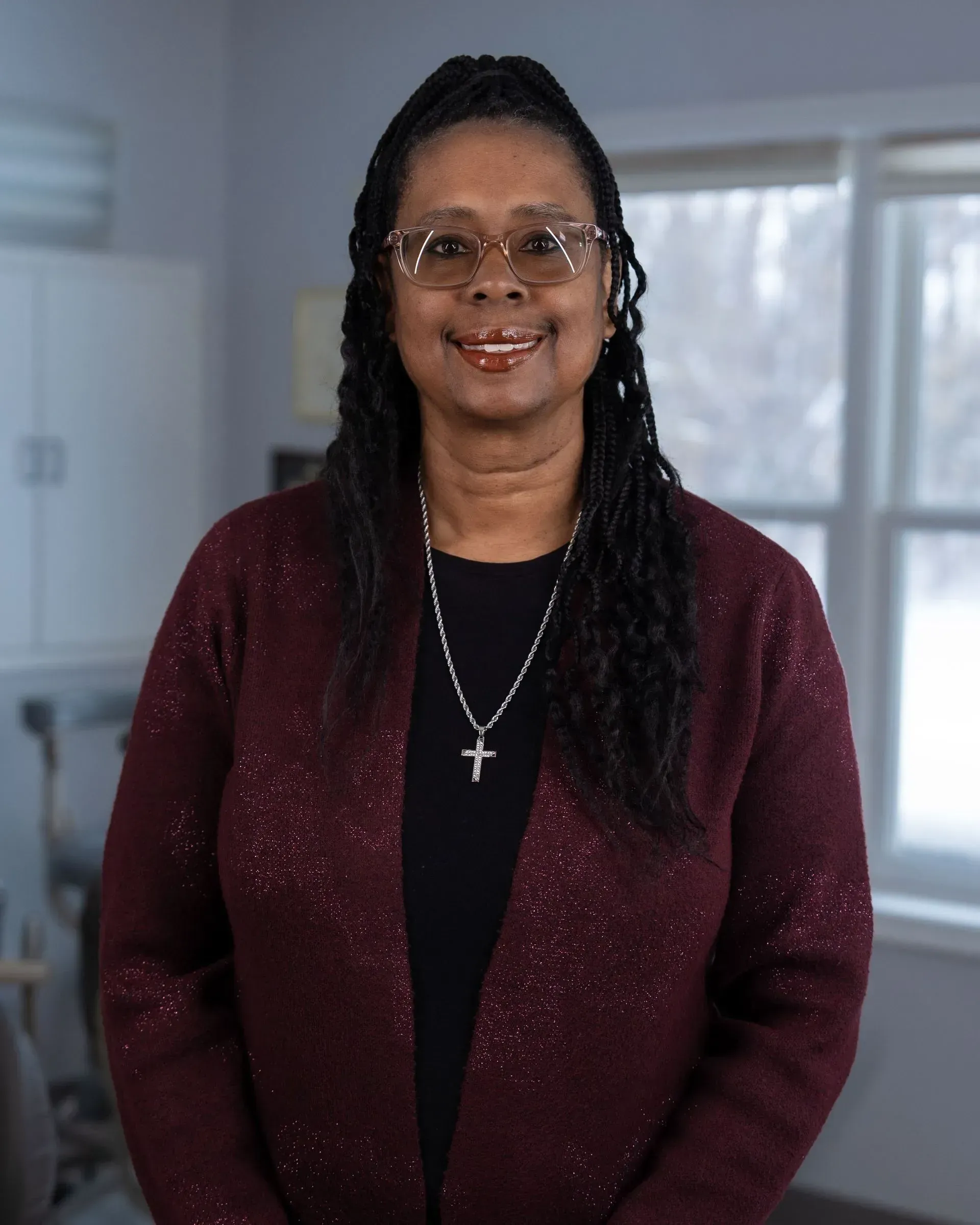Woman in maroon cardigan standing indoors by a bright window, smiling at the camera.