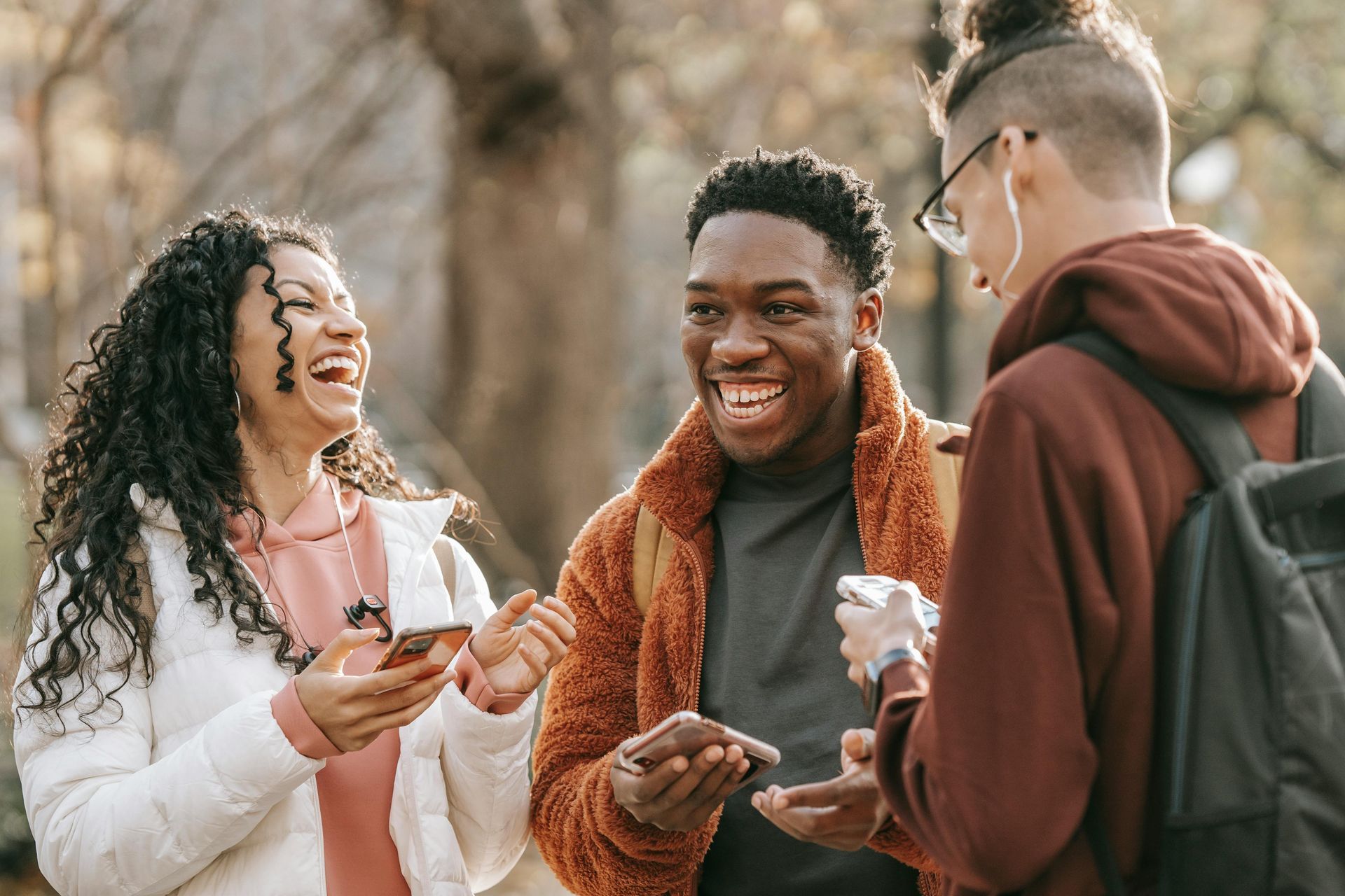 Three young people laughing outdoors. The woman is holding a phone. The men are also holding phones.