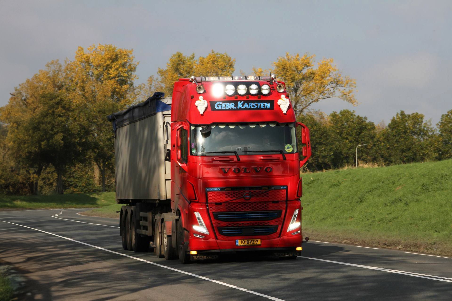 Rode Volvo-vrachtwagen met aanhanger op de weg. Herfstbladeren en een grasheuvel op de achtergrond.