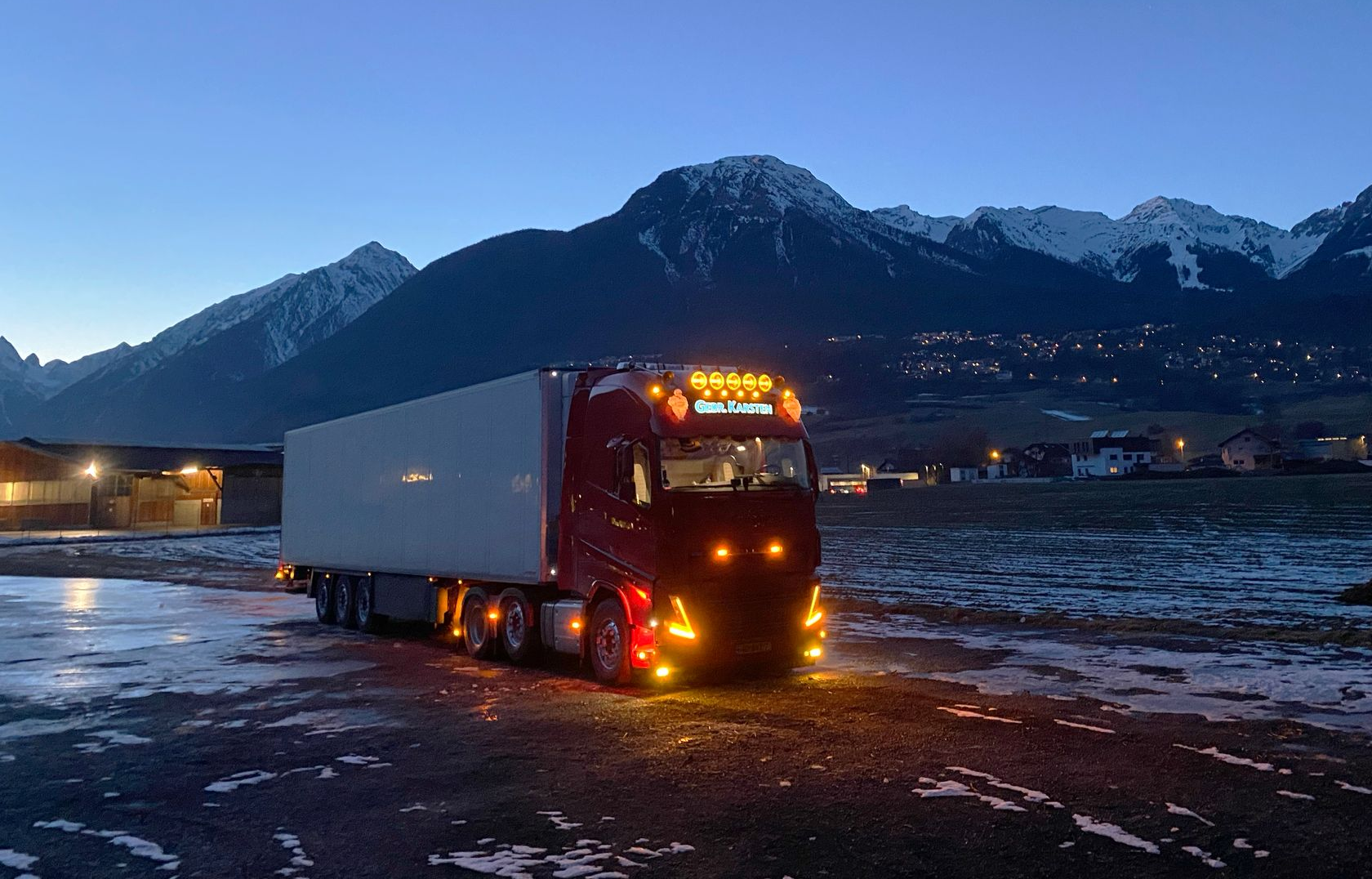 Rode vrachtwagen met aanhanger op een besneeuwd terrein, met bergen op de achtergrond in de schemering.