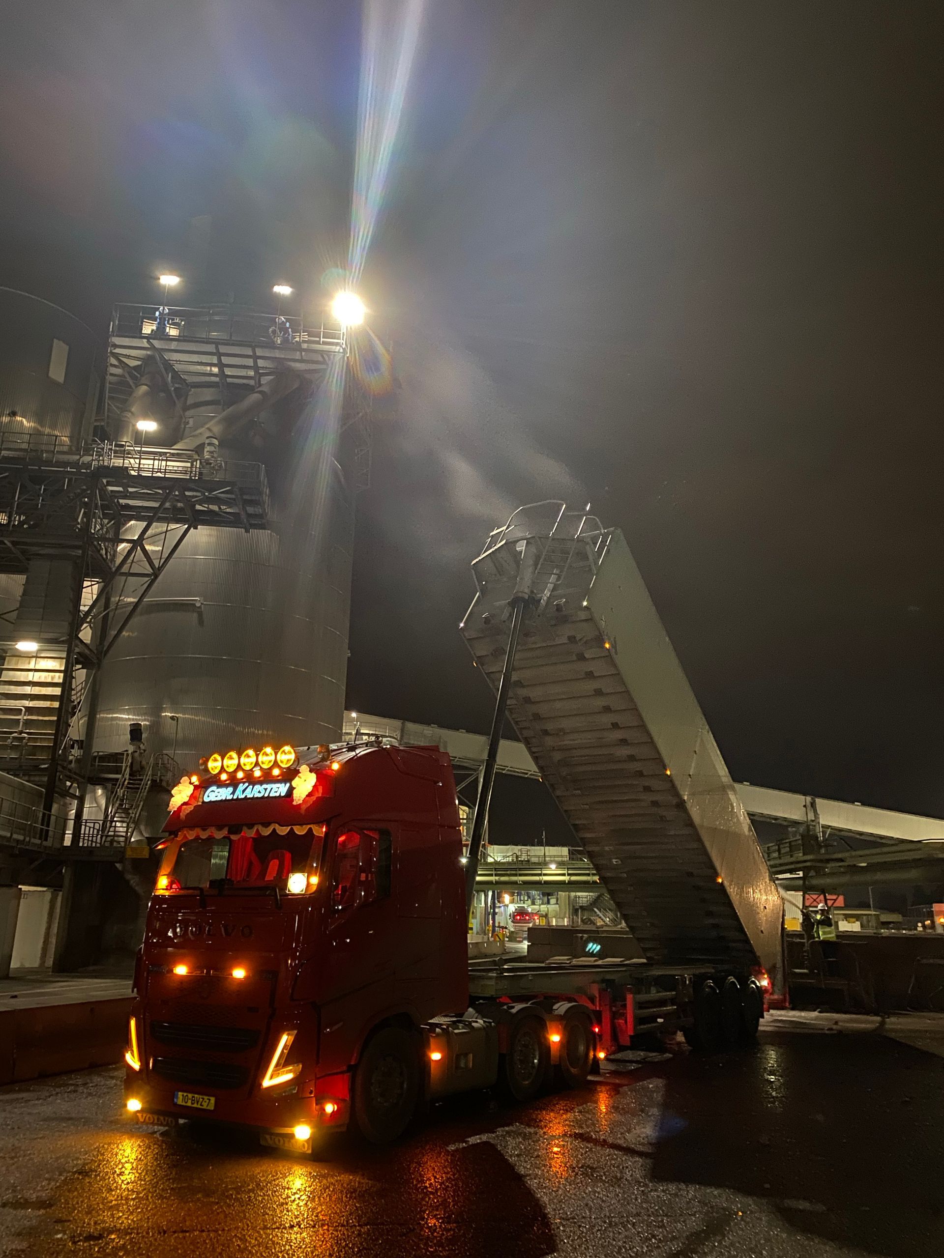 Rode vrachtwagen onder een felle lamp in de nacht, die materiaal in een hoge constructie laadt.