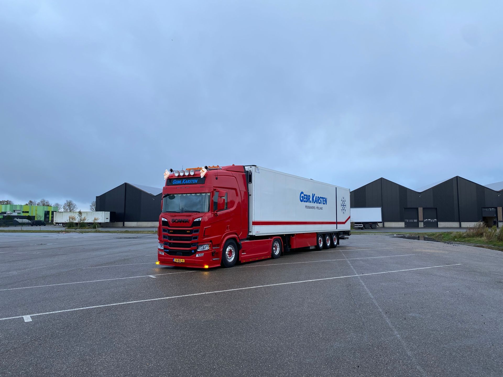 Rood-witte vrachtwagen met aanhanger op een lege parkeerplaats, met een bewolkte lucht op de achtergrond.