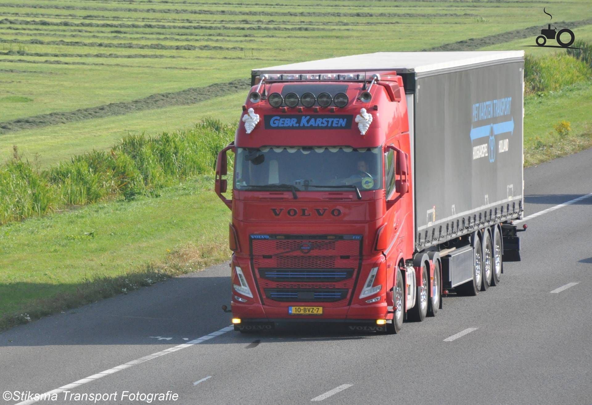 Een rode Volvo-vrachtwagen rijdt over de snelweg met een aanhanger. Groene velden op de achtergrond.