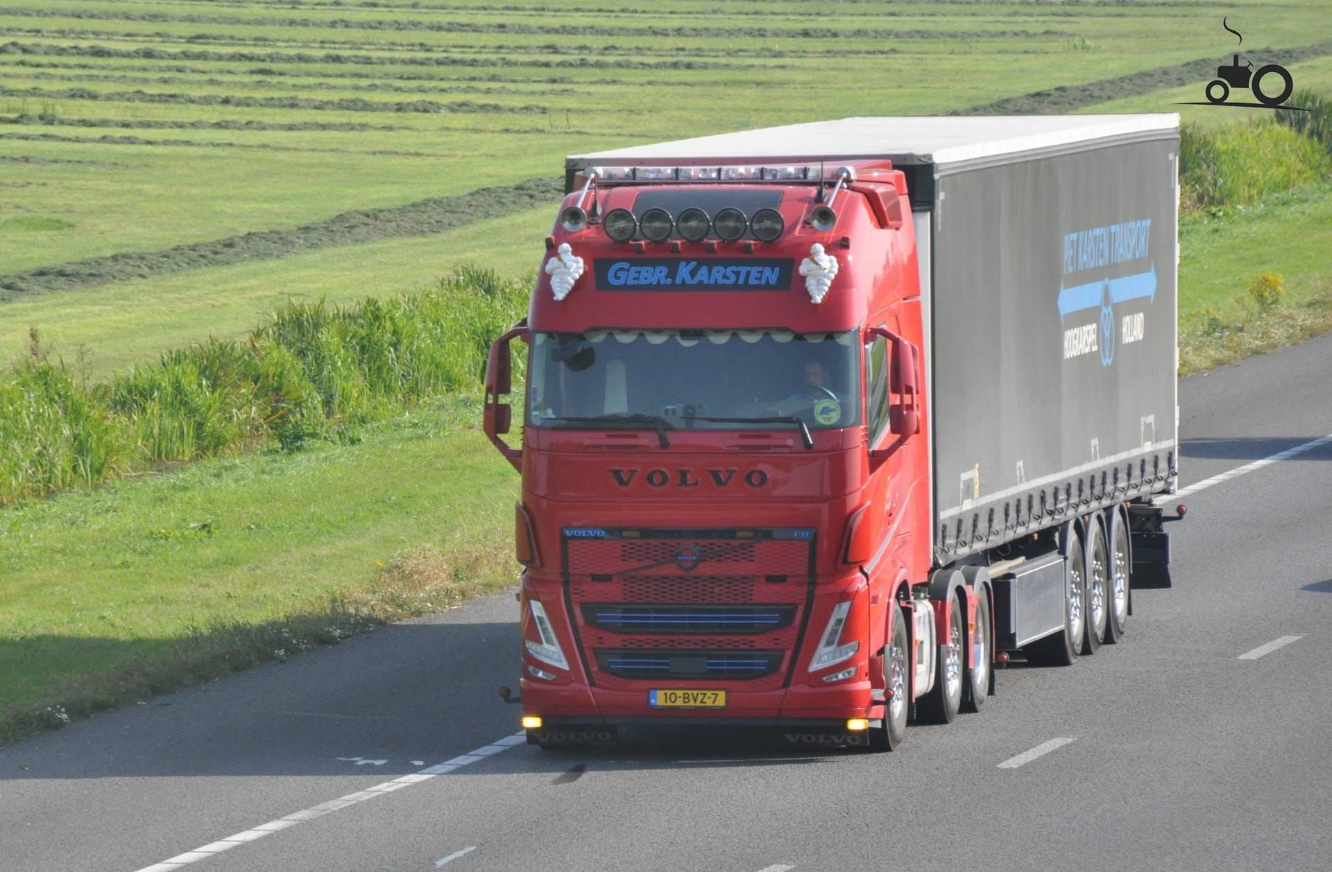 Rode Volvo vrachtwagen met oplegger rijdt op een snelweg, met groene velden op de achtergrond.