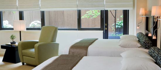A hotel room with two beds, a green armchair, and a view of an outdoor patio through the windows.