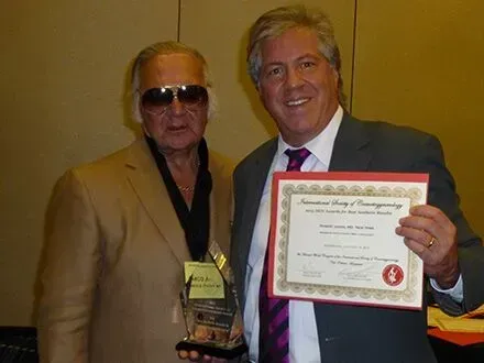 Dr Jason holding an award and a certificate. One wears sunglasses, the other a suit and tie.