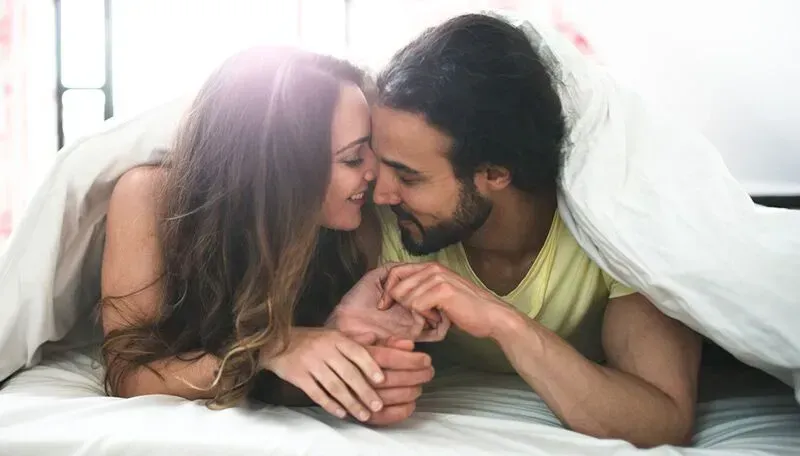Couple in bed, smiling and touching foreheads under a white blanket.