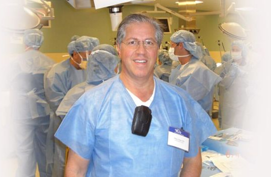 Doctor Robert Jason in blue scrubs smiles in an operating room, with staff in protective gear in the background.