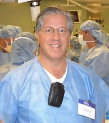 Smiling person in surgical scrubs and glasses in an operating room, name tag visible.