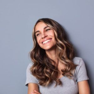 Woman with wavy brown hair smiles broadly, looking up, against a gray background.
