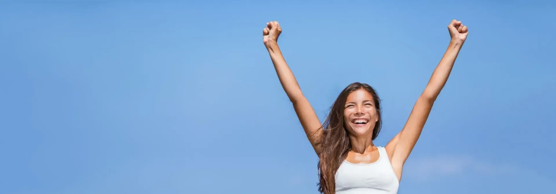 Woman with arms raised, smiling against a clear blue sky.