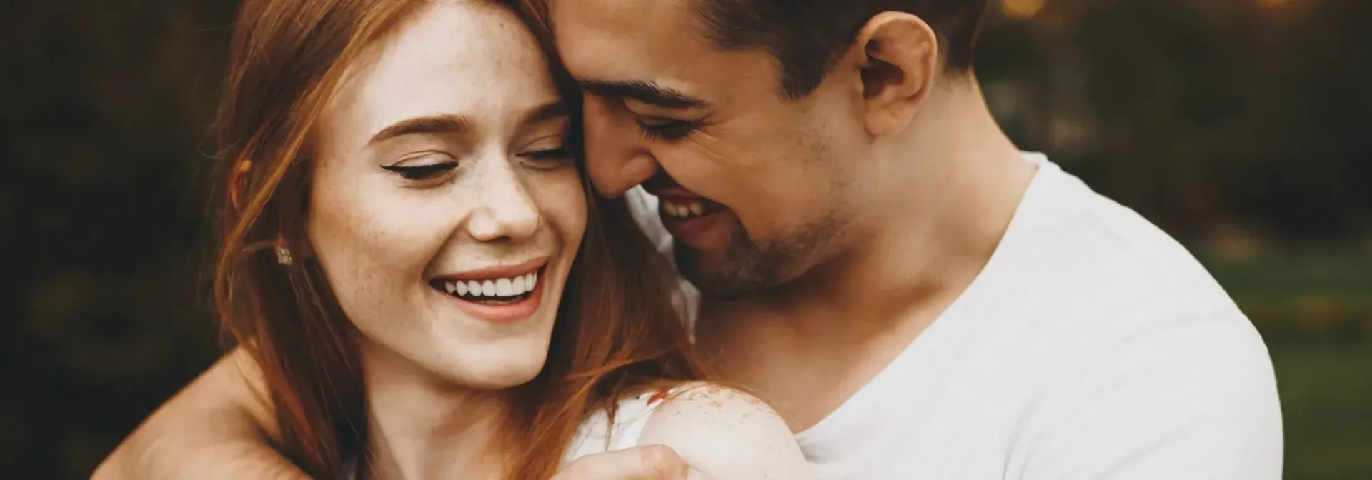 A couple embraces, smiling. The woman has red hair, and the man wears a white shirt. They're outdoors.