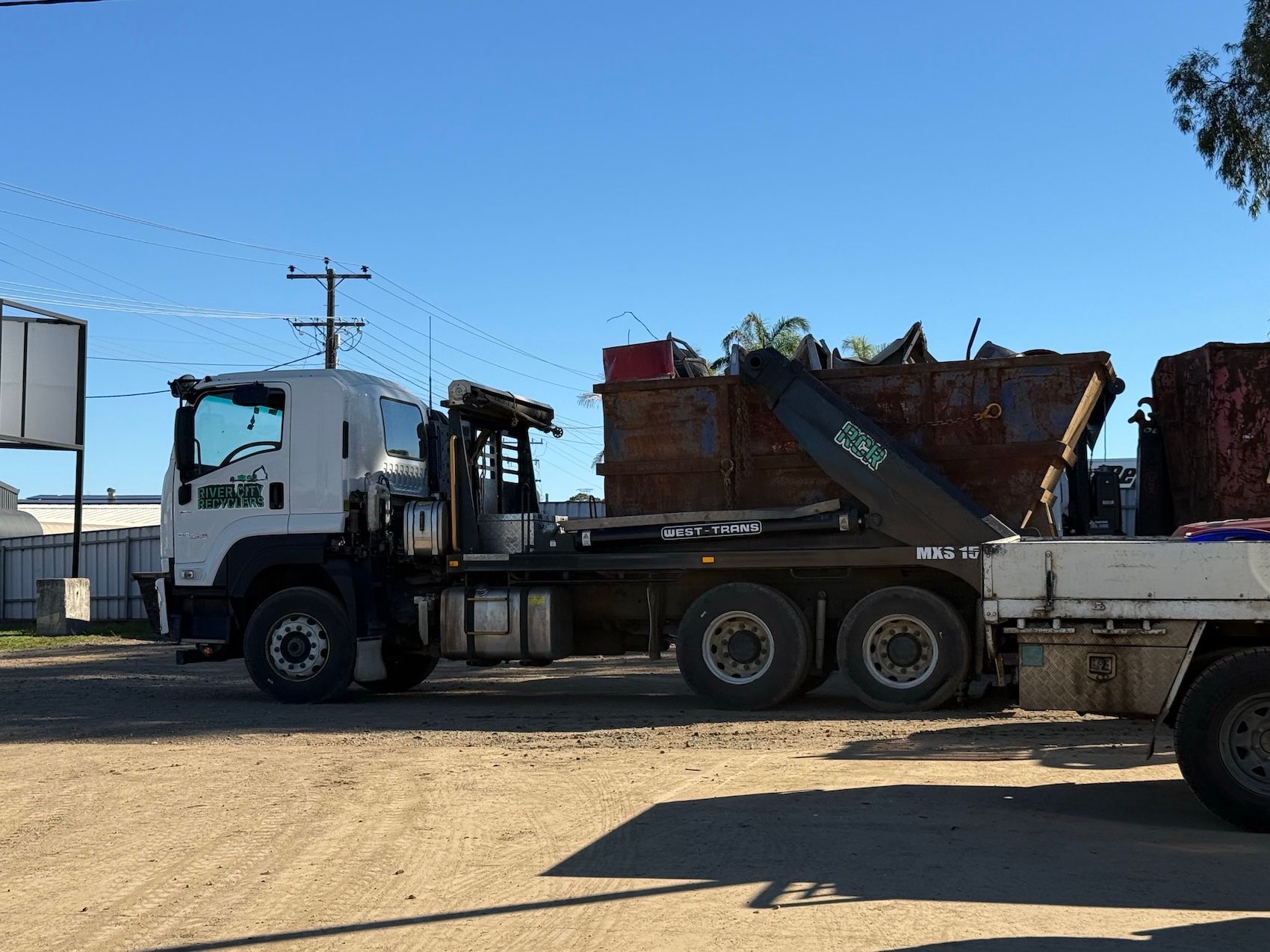White flatbed truck carrying a rusty dumpster on a sunny industrial lot