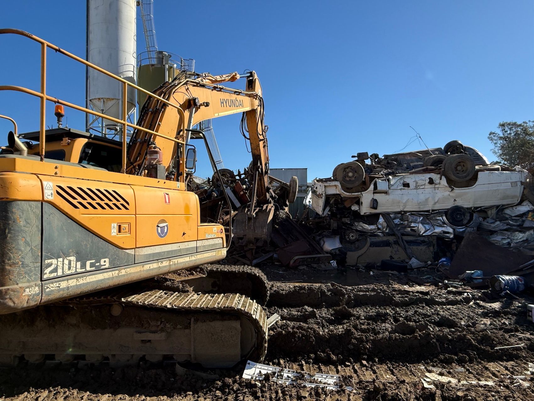 Orange excavator working in a muddy demolition site beside a pile of wrecked cars under a blue sky